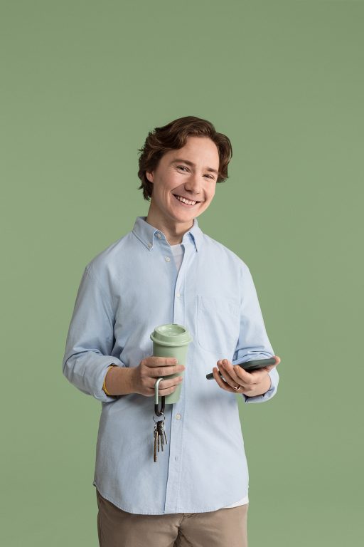 Nuori mies hymyilee kameralle kännykkä ja kahvimuki käsissään.