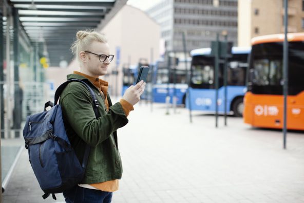 Mies seisoo reppu selässä bussiasemalla ja selaa kännykkää.