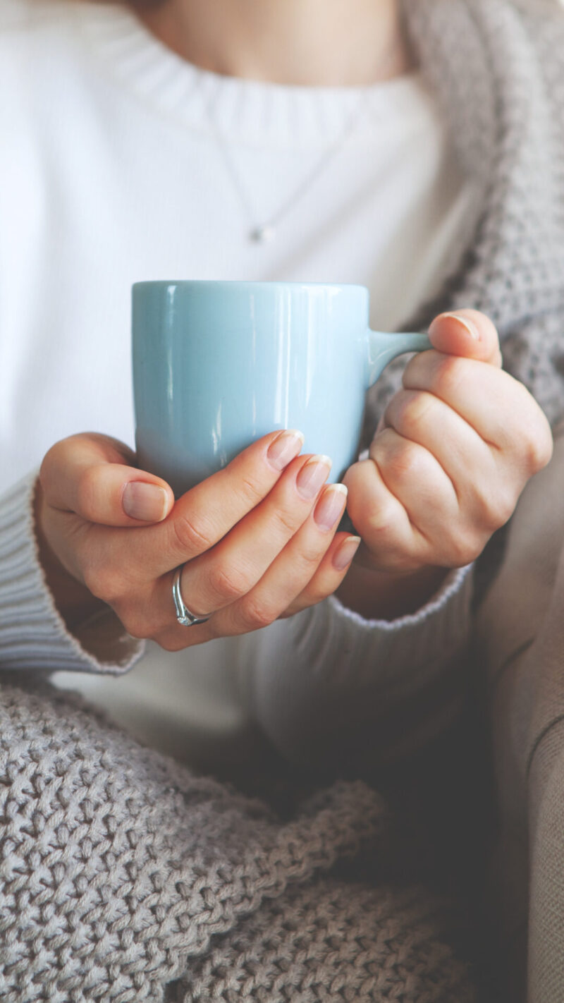 Nainen istuu levollisesti sohvalla viltin alla ja pitää lämmintä juomakuppi käsissään.